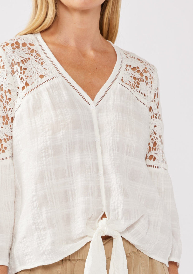 [Color: Off White] A detail image of a blonde model wearing a textured seersucker plaid top with a lace contrast yoke. With a v-neckline, button front, long bell sleeves, and a tie front detail at the hem. A resort top perfect for pairing with denim shorts or any relaxed bottom. 