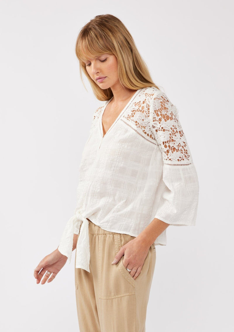 [Color: Off White] A side facing image of a blonde model wearing a textured seersucker plaid top with a lace contrast yoke. With a v-neckline, button front, long bell sleeves, and a tie front detail at the hem. A resort top perfect for pairing with denim shorts or any relaxed bottom. 