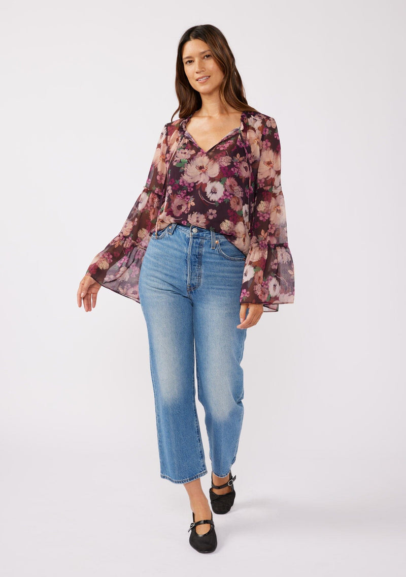 [Color: Plum/Dusty Rose] A full body front-facing image of a brunette model wearing a sheer purple floral chiffon blouse. The blouse features tiered long bell sleeves and a ruffled split v-neckline with tie details. Perfect for day-to-night outings and special occasions in the Fall season. 