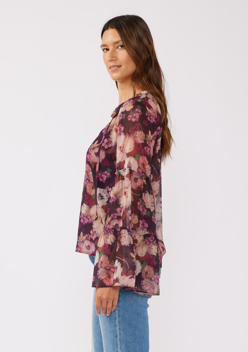 [Color: Plum/Dusty Rose] A side facing image of a brunette model wearing a sheer purple floral chiffon blouse. The blouse features tiered long bell sleeves and a ruffled split v-neckline with tie details. Perfect for day-to-night outings and special occasions in the Fall season. 