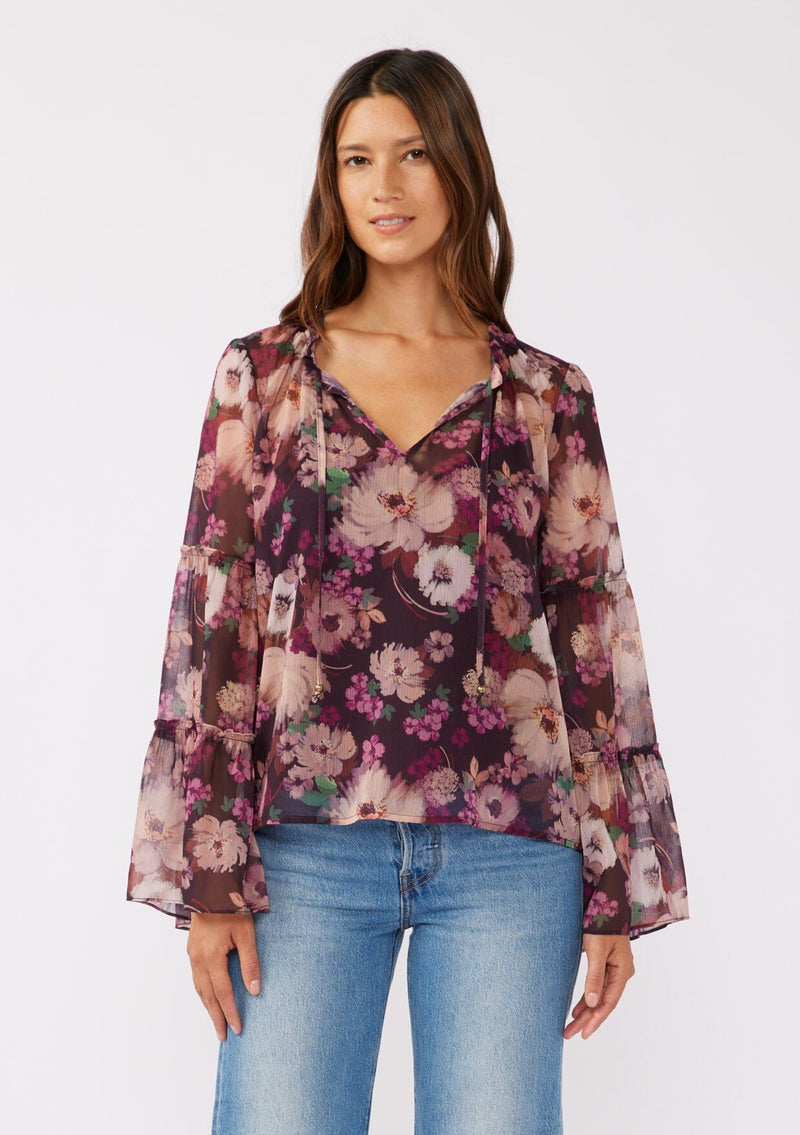 [Color: Plum/Dusty Rose] A front-facing image of a brunette model wearing a sheer purple floral chiffon blouse. The blouse features tiered long bell sleeves and a ruffled split v-neckline with tie details. Perfect for day-to-night outings and special occasions in the Fall season. 