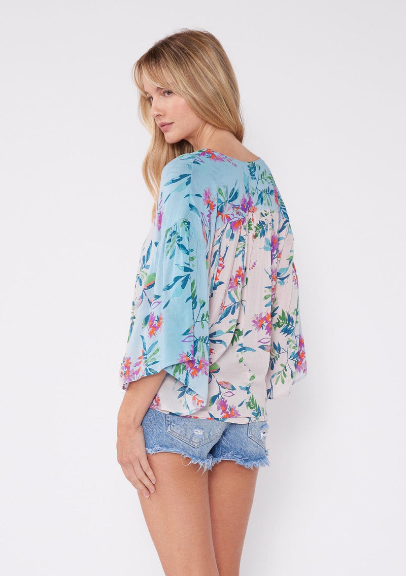 [Color: Light Pink/Purple] A blonde model wearing a vibrant tropical floral print top. This summer top features an ombre effect, surplice v neckline, front tie closure, front elastic hem, and long flowy wide sleeves. A vacation top paired with denim shorts.