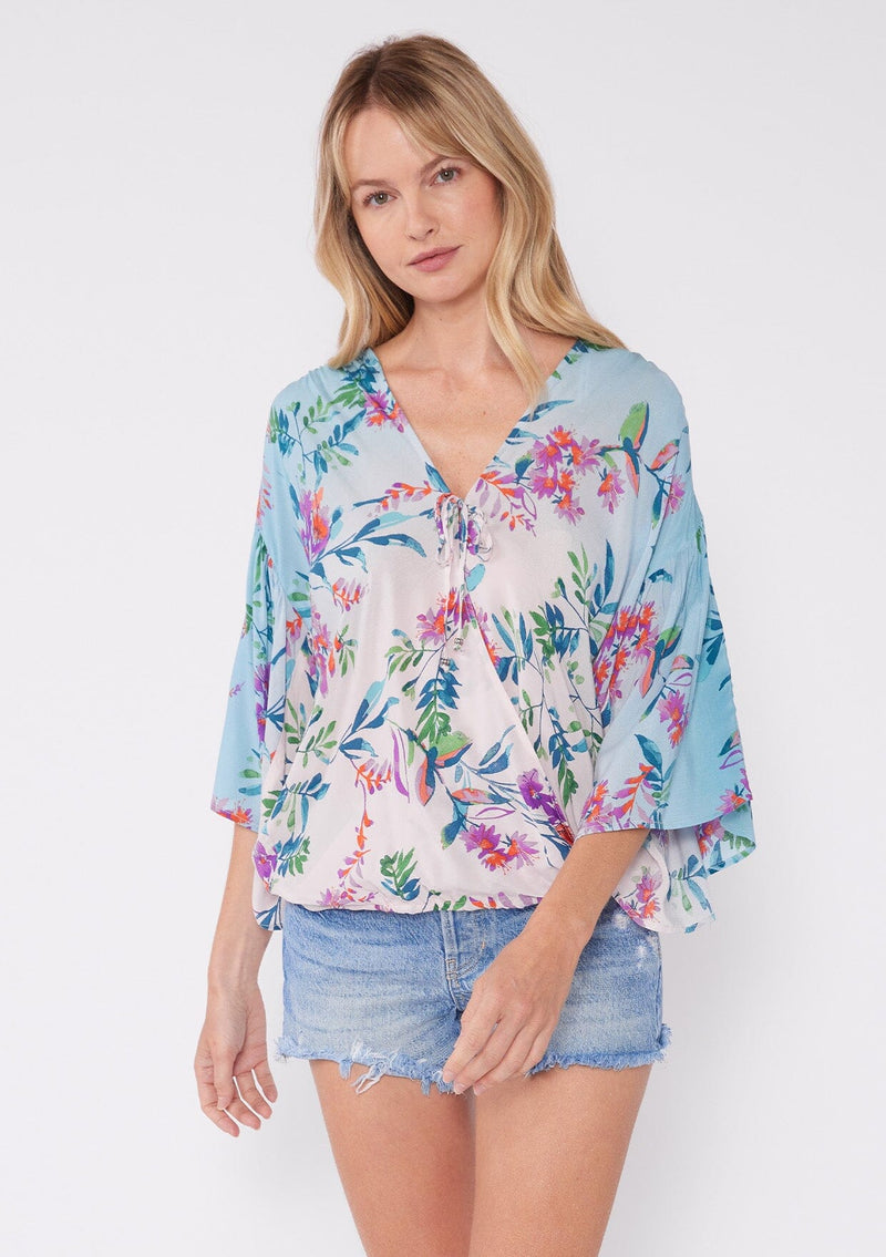 [Color: Light Pink/Purple] A blonde model wearing a vibrant tropical floral print top. This summer top features an ombre effect, surplice v neckline, front tie closure, front elastic hem, and long flowy wide sleeves. A vacation top paired with denim shorts.