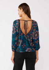 [Color: Teal/Rust] A brunette model wearing an ultra-flowy boho blouse in a teal floral print with contrast piping. Featuring a wide v neckline, long sleeves, and an open back with tie detail.