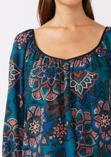 [Color: Teal/Rust] A brunette model wearing an ultra-flowy boho blouse in a teal floral print with contrast piping. Featuring a wide v neckline, long sleeves, and an open back with tie detail.