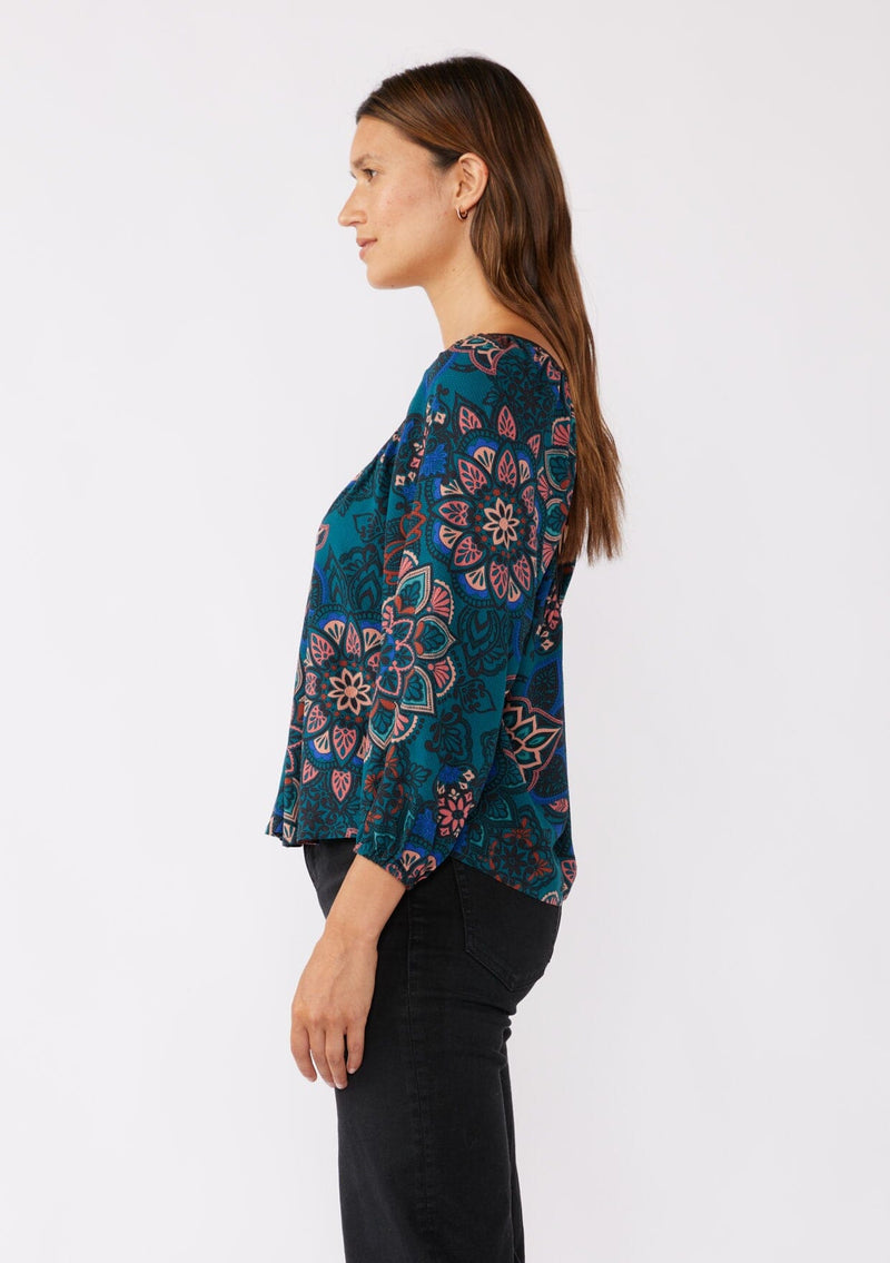 [Color: Teal/Rust] A brunette model wearing an ultra-flowy boho blouse in a teal floral print with contrast piping. Featuring a wide v neckline, long sleeves, and an open back with tie detail.