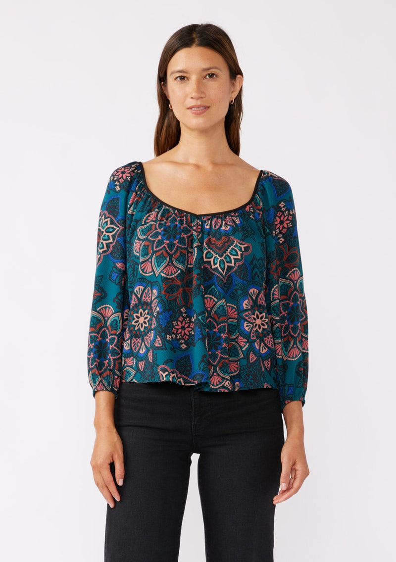 [Color: Teal/Rust] A brunette model wearing an ultra-flowy boho blouse in a teal floral print with contrast piping. Featuring a wide v neckline, long sleeves, and an open back with tie detail.  