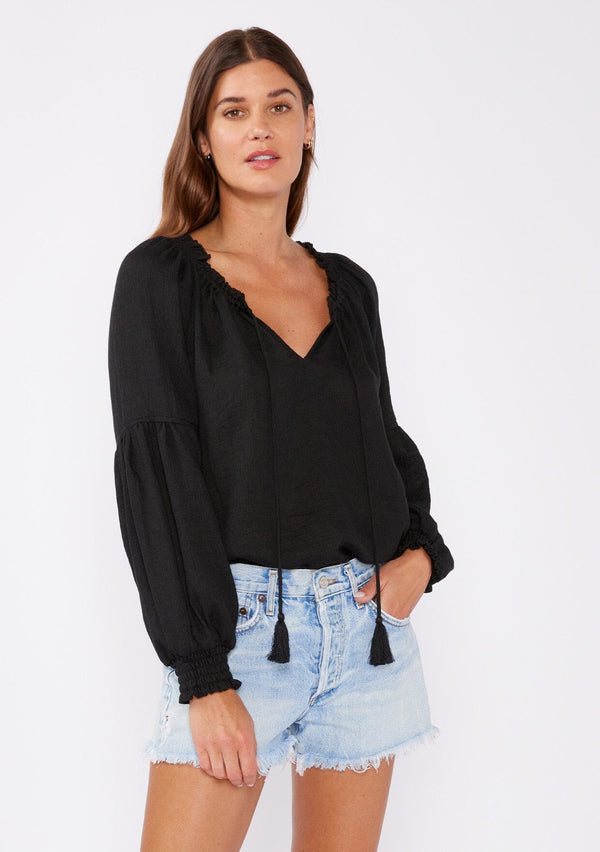 [Color: Black] A brunette model wearing a classic lightweight sheer black peasant top. With a ruffled neckline, tassel ties, long voluminous bishop sleeves, and a relaxed flowy fit.