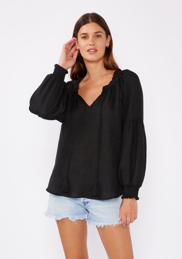 [Color: Black] A brunette model wearing a classic lightweight sheer black peasant top. With a ruffled neckline, tassel ties, long voluminous bishop sleeves, and a relaxed flowy fit.