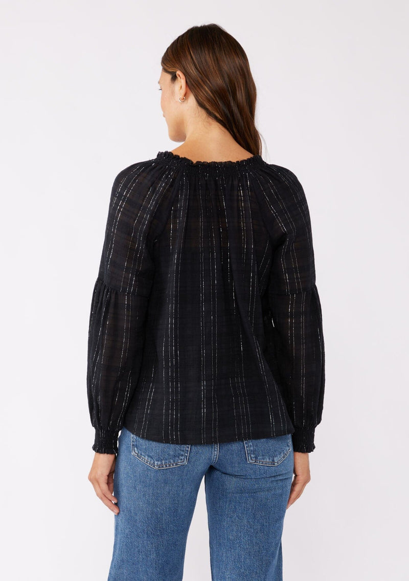 [Color: Black] A brunette model wearing a black peasant top in a jacquard stripe with metallic lurex details. Featuring a classic split neckline with tie detail, long sleeves, smocked cuffs, and a flowy, relaxed silhouette. An elevated casual peasant top, perfect for any holiday celebration or nice dinners.