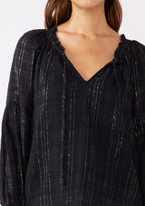 [Color: Black] A brunette model wearing a black peasant top in a jacquard stripe with metallic lurex details. Featuring a classic split neckline with tie detail, long sleeves, smocked cuffs, and a flowy, relaxed silhouette. An elevated casual peasant top, perfect for any holiday celebration or nice dinners.