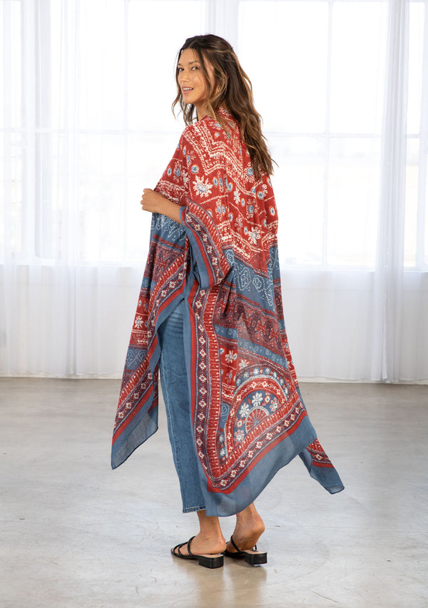 [Color: Blue/Brick] A model wearing a red and blue bohemian mid length kimono. Featuring half length kimono sleeves, side slits, an open front, and a multi colored bohemian pattern throughout.