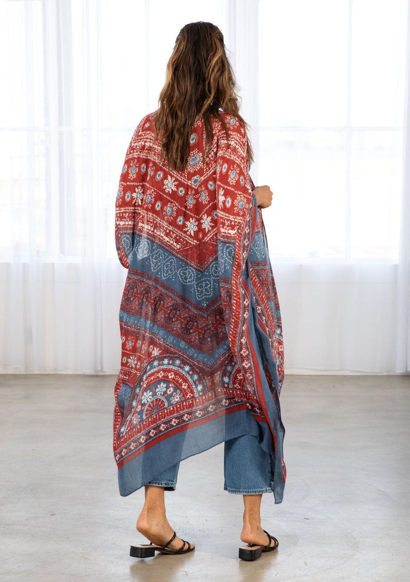 [Color: Blue/Brick] A model wearing a red and blue bohemian mid length kimono. Featuring half length kimono sleeves, side slits, an open front, and a multi colored bohemian pattern throughout.