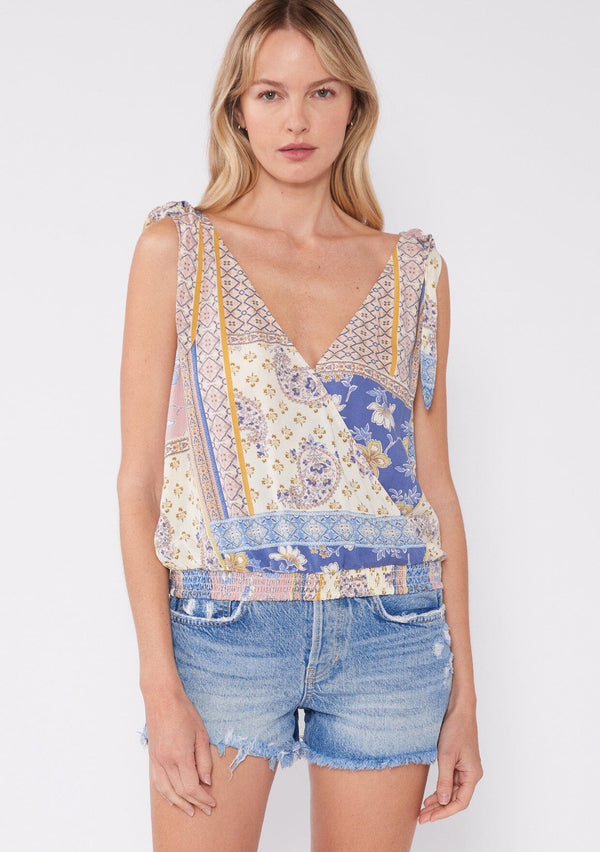 [Color: Ivory/Lilac] A front facing image of a blonde model wearing a bohemian tank top in an ivory and lilac purple patchwork floral print. With tie shoulder details, a deep surplice v neckline, and a smocked elastic waist. 