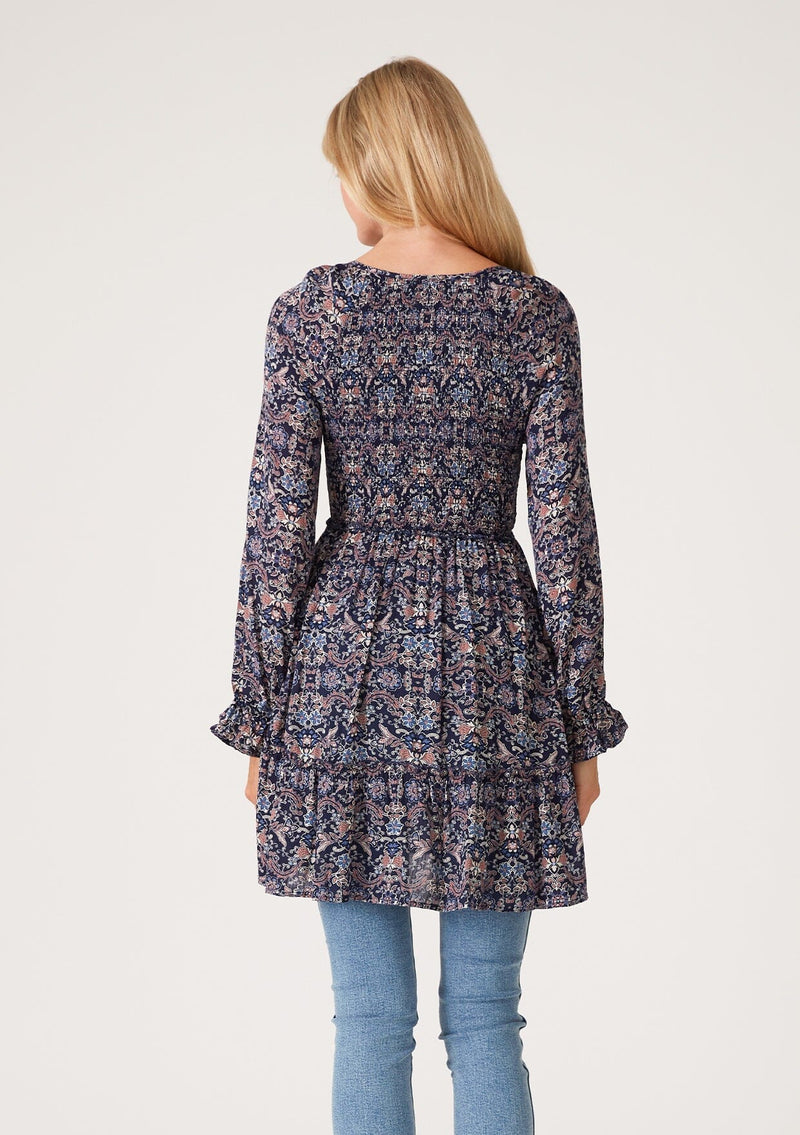 [Color: Navy/Tan] A back facing image of a blonde model wearing a navy blue and tan paisley print tunic top. A vintage inspired top with long sleeves, a smocked bodice, an empire waist, and a deep v neckline.