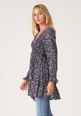 [Color: Navy/Tan] A side facing image of a blonde model wearing a navy blue and tan paisley print tunic top. A vintage inspired top with long sleeves, a smocked bodice, an empire waist, and a deep v neckline.