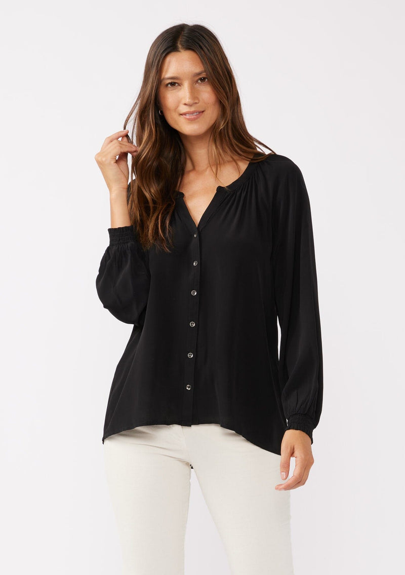 [Color: Black] A front facing image of a brunette model wearing a soft and silky black button front blouse with long sleeves, smocked wrist cuffs, and a split v neckline.