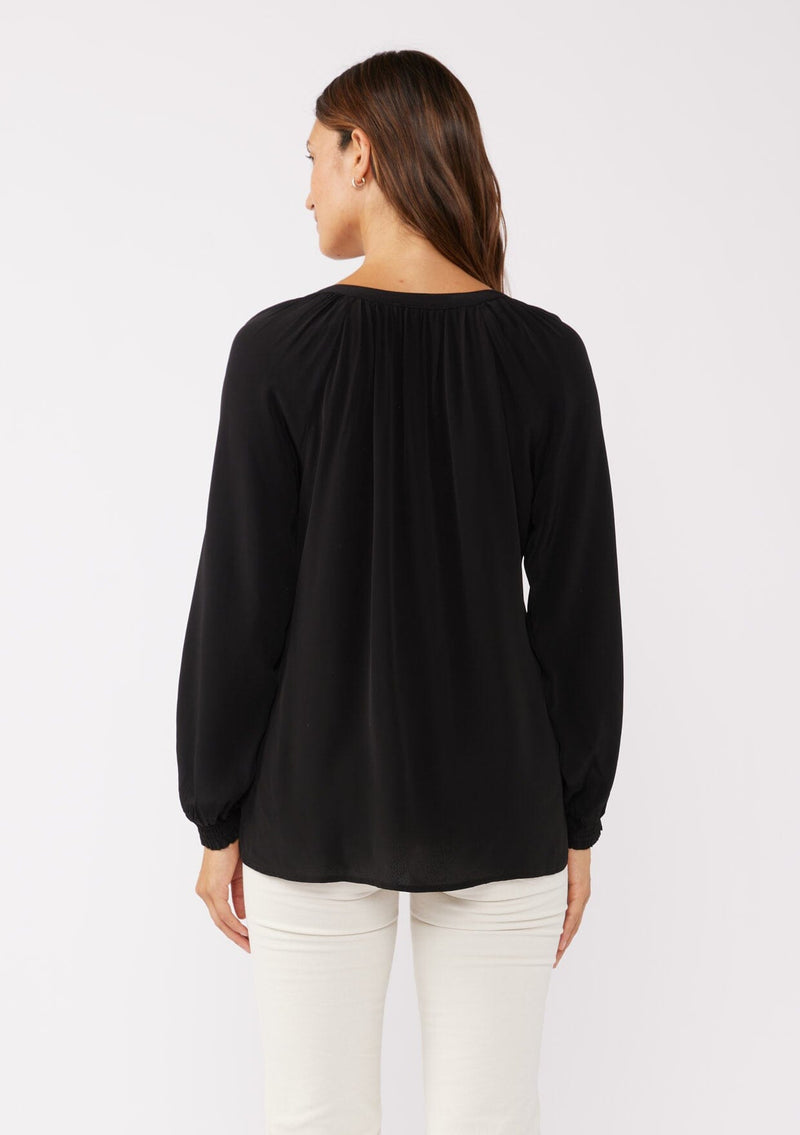 [Color: Black] A back facing image of a brunette model wearing a soft and silky black button front blouse with long sleeves, smocked wrist cuffs, and a split v neckline.