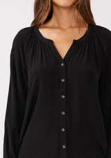 [Color: Black] A detailed image of a brunette model wearing a soft and silky black button front blouse with long sleeves, smocked wrist cuffs, and a split v neckline.