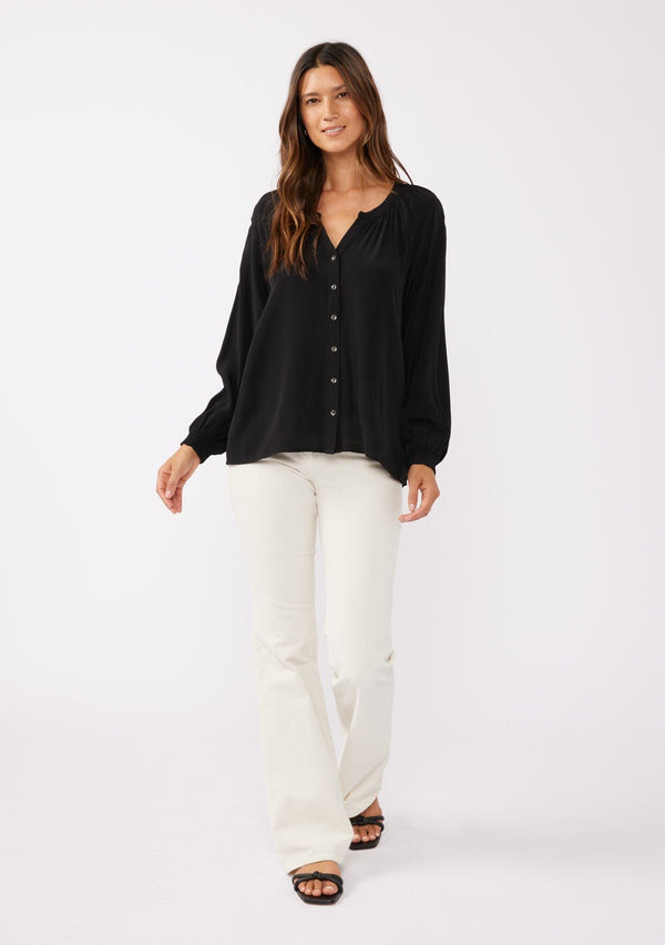 [Color: Black] A full body front facing image of a brunette model wearing a soft and silky black button front blouse with long sleeves, smocked wrist cuffs, and a split v neckline.