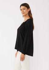 [Color: Black] A side facing image of a brunette model wearing a soft and silky black button front blouse with long sleeves, smocked wrist cuffs, and a split v neckline.