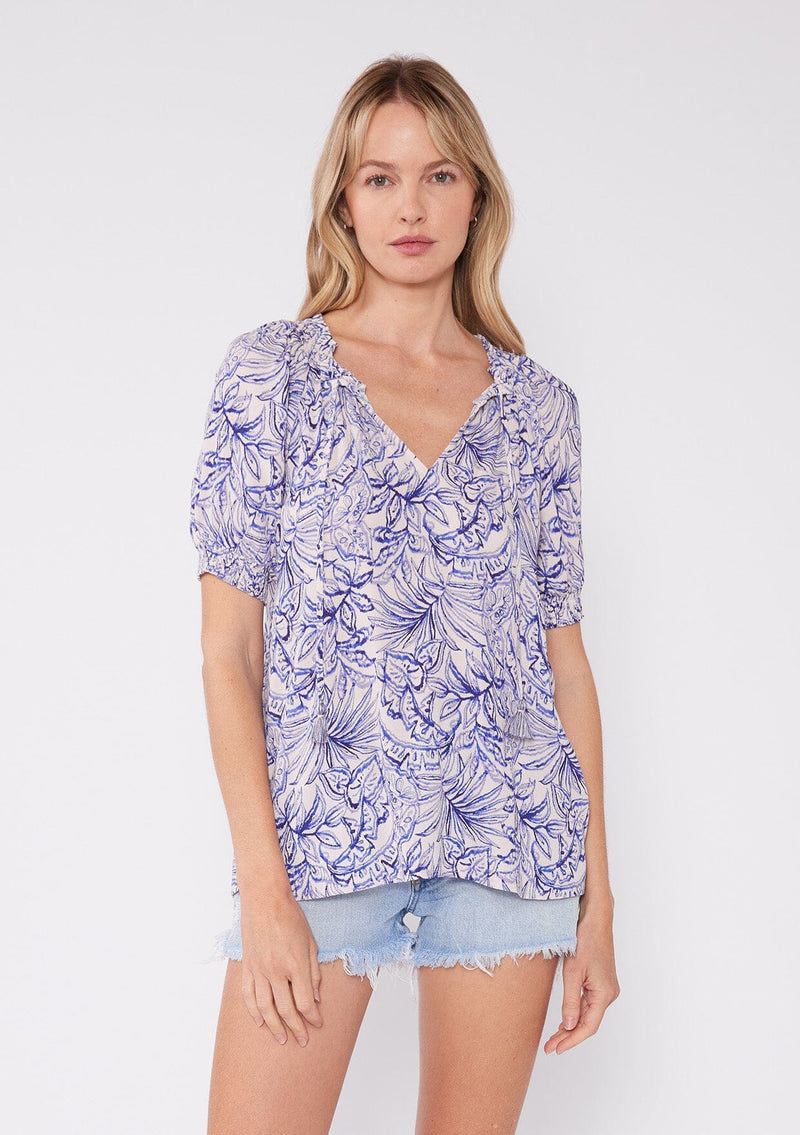 [Color: Bone/Lilac] Beautiful image of short sleeve white bohemian blouse with purple botanical print and a split v-neckline with tassel ties. A cute summer blouse with a relaxed fit.