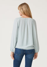 [Color: Sky] A back facing image of a blonde model wearing a soft and silky light blue bohemian blouse with voluminous long sleeves, a plunging surplice v neckline, and an elastic front waist.