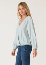 [Color: Sky] A side facing image of a blonde model wearing a soft and silky light blue bohemian blouse with voluminous long sleeves, a plunging surplice v neckline, and an elastic front waist.