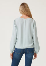 [Color: Sky] A back facing image of a blonde model wearing a soft and silky light blue crepe long sleeve top with a tie front detail. 