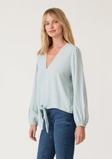 [Color: Sky] A side facing image of a blonde model wearing a soft and silky light blue crepe long sleeve top with a tie front detail. 