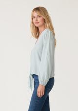 [Color: Sky] A side facing image of a blonde model wearing a soft and silky light blue crepe long sleeve top with a tie front detail. 