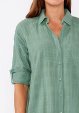 [Color: Dusty Olive] Brunette woman wearing an olive green plaid seersucker shirt with a button front, roll tab long sleeve, collared neckline. A great top to pair with denim pants or shorts.