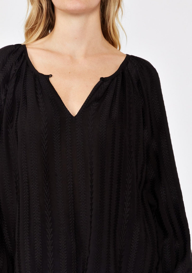 [Color: Black] A detailed image of a blonde model wearing a bohemian black blouse with embroidered details. With long raglan sleeves, a round neckline with a single button closure, a front keyhole, and a relaxed fit.