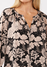 [Color: Black/Taupe] A detail image of a blonde model wearing a black and taupe floral print blouse. This boho casual blouse features a self covered button front, a split v-neckline, and 3/4 length sleeves. Perfect for any casual outing or vacation. 