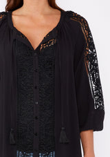 [Color: Black] A brunette model wearing a black bohemian resort blouse with voluminous long sleeves, a button front, tassel neck ties, and lace trim throughout.