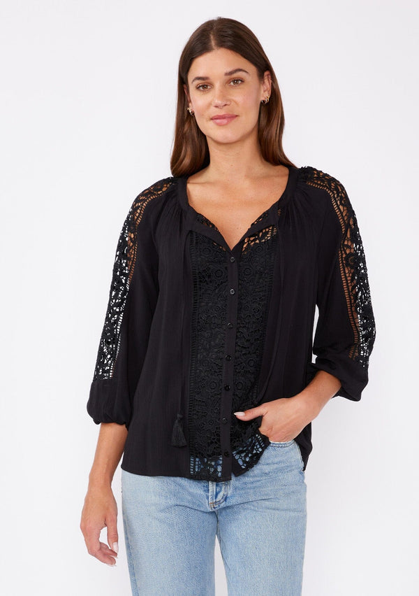 [Color: Black] A brunette model wearing a black bohemian resort blouse with voluminous long sleeves, a button front, tassel neck ties, and lace trim throughout.