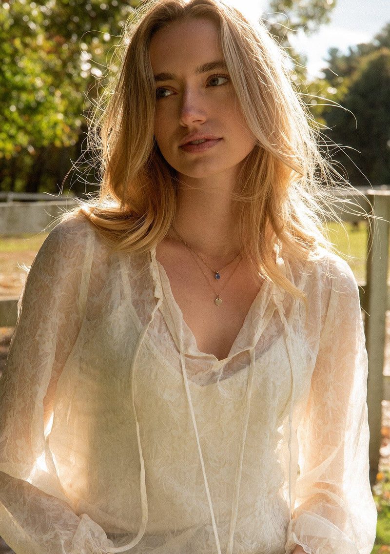 [Color: Ivory] A detail image of a blonde model wearing a crinkled sheer chiffon blouse with a floral pattern in ivory off white. This relaxed fit blouse features a double tie neck detail, long sleeves with ruffled elastic cuffs, and a detachable cami. 