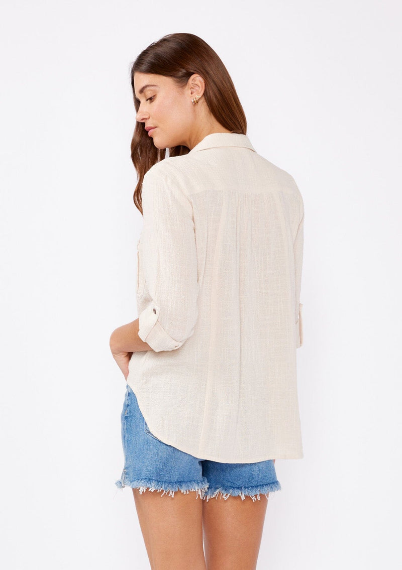 [Color: Sand] An image of fa brunette model wearing a ivory light beige cotton shirt. With long rolled sleeved, a button tab sleeve closure, a button front, two front patch pockets, a tie front waist detail, and a high low hemline. Styled with cut off denim for the summer season.