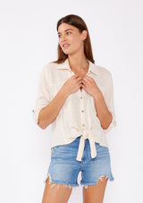 [Color: Sand] An image of fa brunette model wearing a ivory light beige cotton shirt. With long rolled sleeved, a button tab sleeve closure, a button front, two front patch pockets, a tie front waist detail, and a high low hemline. Styled with cut off denim for the summer season.