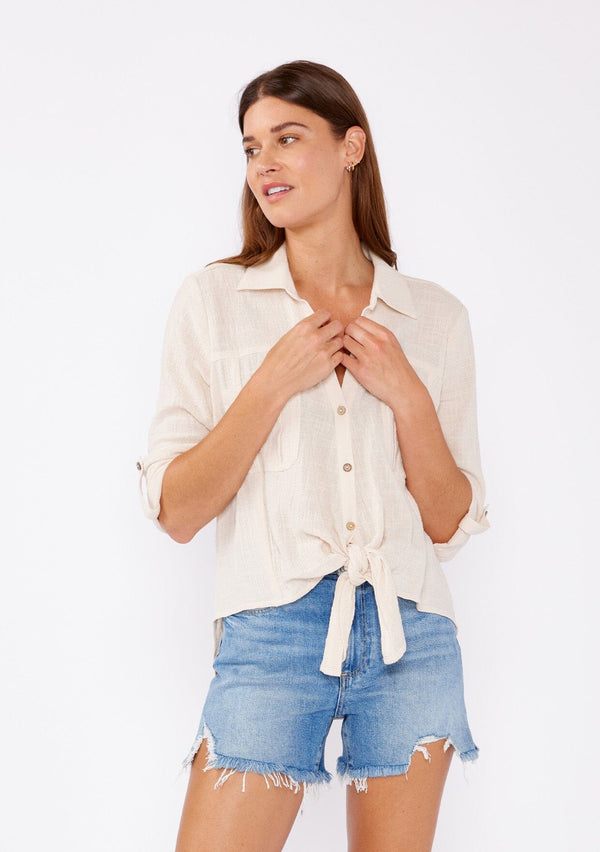[Color: Sand] An image of fa brunette model wearing a ivory light beige cotton shirt. With long rolled sleeved, a button tab sleeve closure, a button front, two front patch pockets, a tie front waist detail, and a high low hemline. Styled with cut off denim for the summer season.