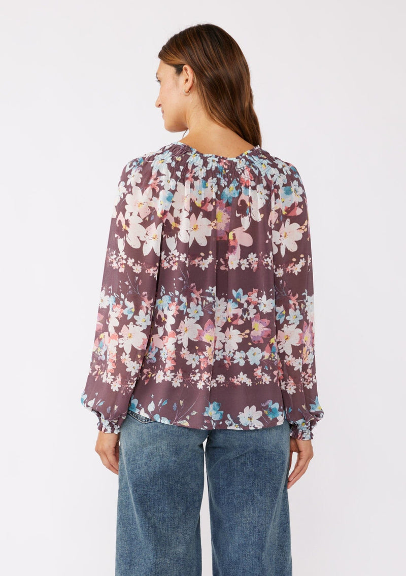 [Color: Plum/Blue] A brunette model wearing an ultra-flowy blouse, crafted in lightweight chiffon fabric with a floral print. This purple blend floral blouse features smocked elastic details, a split v neckline, and a tie detail at the neck. A relaxed fit top for the fall season. 