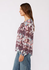 [Color: Plum/Blue] A brunette model wearing an ultra-flowy blouse, crafted in lightweight chiffon fabric with a floral print. This purple blend floral blouse features smocked elastic details, a split v neckline, and a tie detail at the neck. A relaxed fit top for the fall season. 