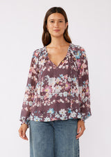 [Color: Plum/Blue] A brunette model wearing an ultra-flowy blouse, crafted in lightweight chiffon fabric with a floral print. This purple blend floral blouse features smocked elastic details, a split v neckline, and a tie detail at the neck. A relaxed fit top for the fall season. 