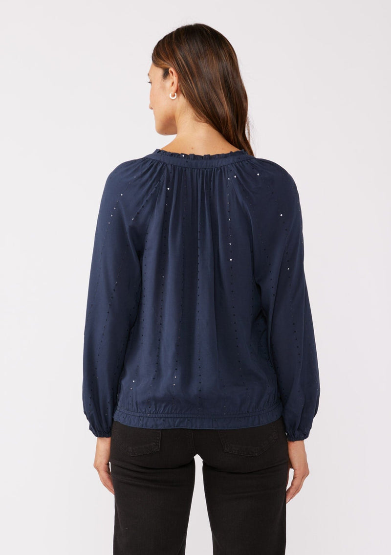 [Color: Midnight] A back image of a brunette model wearing a sparkly sequin stripe top in navy blue. Featuring a split v neckline with tassel ties, long sleeves, and a comfortable elastic waistline. Styled dark denim for the fall season.