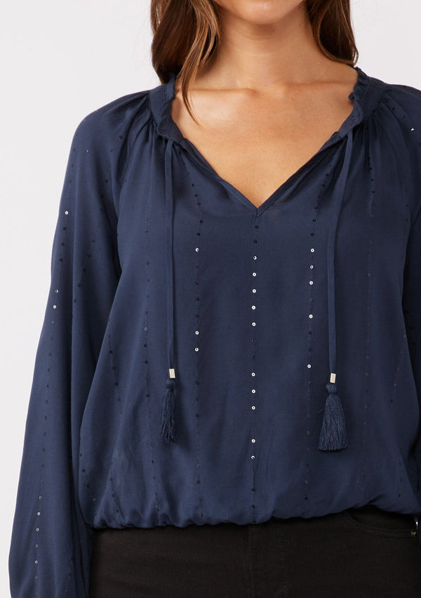 [Color: Midnight] A detailed image of a brunette model wearing a sparkly sequin stripe top in navy blue. Featuring a split v neckline with tassel ties, long sleeves, and a comfortable elastic waistline. Styled dark denim for the fall season.