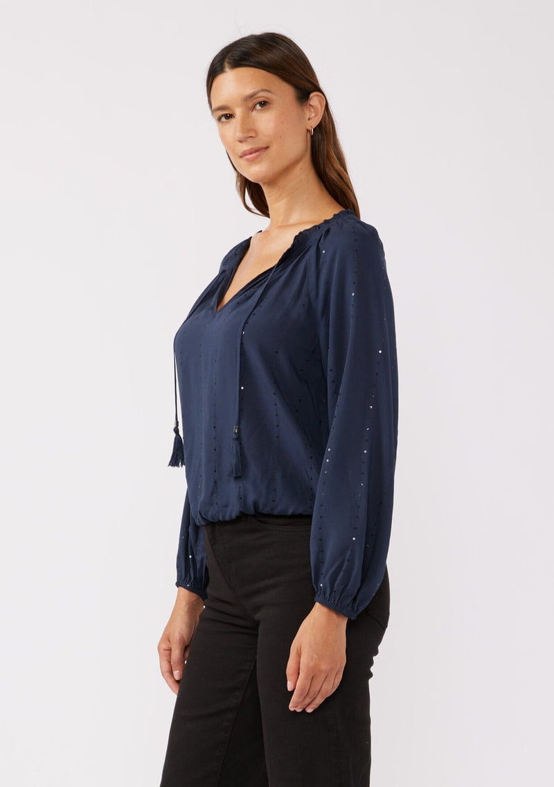 [Color: Midnight] A side image of a brunette model wearing a sparkly sequin stripe top in navy blue. Featuring a split v neckline with tassel ties, long sleeves, and a comfortable elastic waistline. Styled dark denim for the fall season.