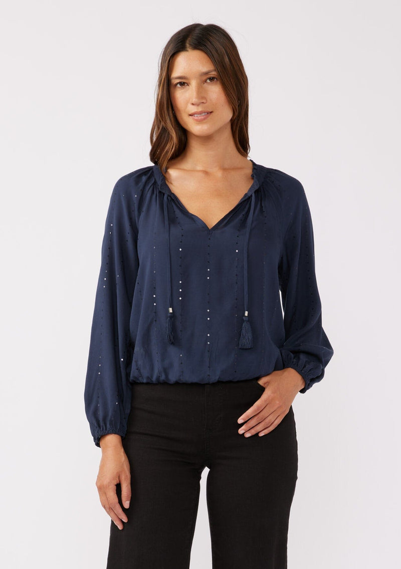 [Color: Midnight] A front facing image of a brunette model wearing a sparkly sequin stripe top in navy blue. Featuring a split v neckline with tassel ties, long sleeves, and a comfortable elastic waistline. Styled dark denim for the fall season.