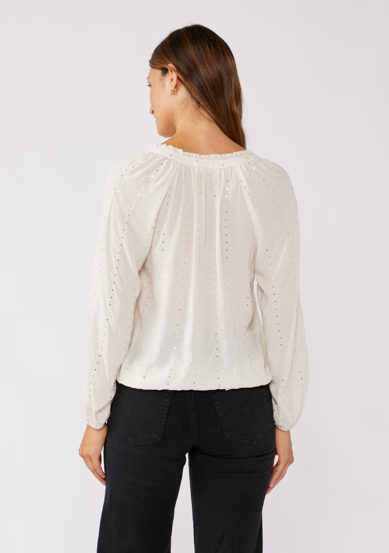 [Color: Natural/Gold] A back facing image of a brunette model wearing a sparkly sequin strip top in ivory off white. Featuring a split v neckline with tassel ties, long sleeves, and a comfortable elastic waistline. Styled for the fall season with black denim.