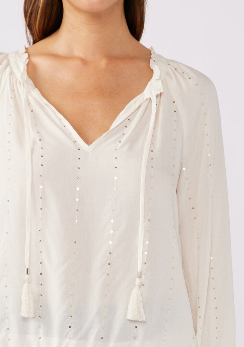 [Color: Natural/Gold] A detailed image of a brunette model wearing a sparkly sequin strip top in ivory off white. Featuring a split v neckline with tassel ties, long sleeves, and a comfortable elastic waistline. 