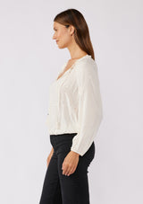 [Color: Natural/Gold] A side facing image of a brunette model wearing a sparkly sequin strip top in ivory off white. Featuring a split v neckline with tassel ties, long sleeves, and a comfortable elastic waistline. Styled for the fall season with black denim jeans.